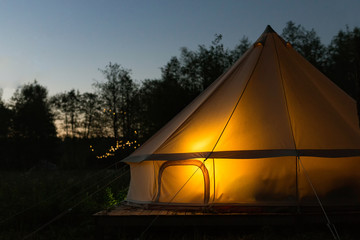 Illuminated yellow camping bell tent at night. Glamping bell tent glows at forest