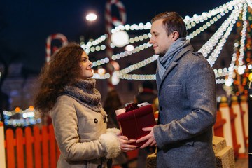 Gift box in the hands of a man and a woman against the background of Christmas street lights. Romantic date Loving couple on Valentine's Day Outdoors. Winter wonderland city  scene. New Year party.