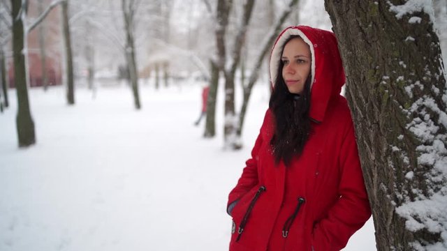 Portrait of young woman in red jacket and hood in winter season. Beautiful lady stands near tree and looks away. Fluffy snow envelops everything around.