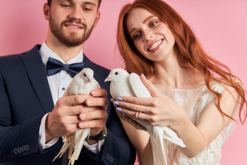 Beautiful caucasian couple after marriage proposal stand holding doves in hands, isolated over pink background