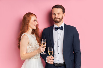 Beautiful woman and handsome na in tux with bow celebrate their wedding day together holding champagne, isolated over pink background