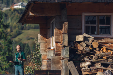 Freelancer bearded man is standing near log house in mountains with laptop and talking on phone.