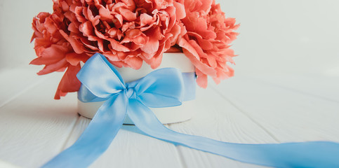 Cropped beautiful red peony flower bouquet in round box with blue sattin ribbon bow on white wooden background. Gift, greeting, compliment idea. Wide banner. Soft selective focus. Copy space.