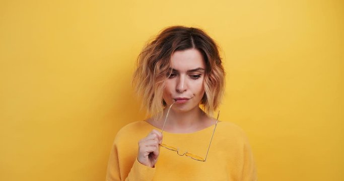Portrait young woman thinks questioningly, holds glasses, finding solution. Idea pointing forward index finger on isolated yellow background, looking directly camera smiling. Monotonous. Emotions