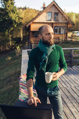 Fototapeta premium freelancer bearded man is standing on platform in mountains with laptop and drinks coffee.