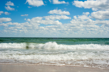 Blue sea wave on sandy beach. Beautiful sea waves on a background of blue sky with clouds on a sunny summer day. Sea waves roll on yellow sand.
