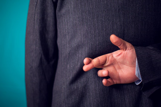 Businessman in suit keeping crossed fingers gesture behind his back in front of black background