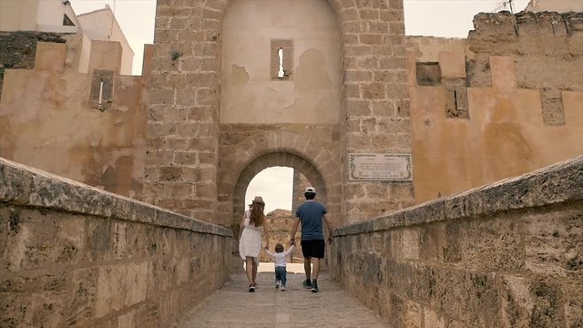 Mother and father are holding their baby boy hands and helping him to make first steps on an old cobbled street at a medieval castle in Spain in Bunyol or Bunol near Valencia. Young family on vacation
