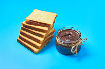 Single glass transparent jar with rope full of melted chocolate or cream near heap of pieces of vanilla bread lies on blue desk on kitchen. Concept of nutritious breakfast or lunch