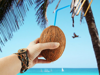 Coconut with straw in female hand with authentic bracelets on background of palm beach, azure sea with sailboats and bird in sky. Time to travel. Rest and relaxation concept.  