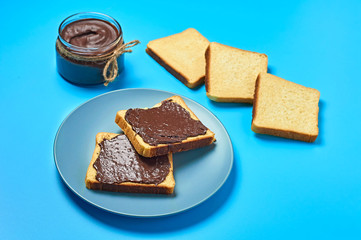 Glass jar full of melted chocolate near plate with pieces of vanilla bread lies on blue desk on kitchen. Concept of nutritious breakfast or lunch