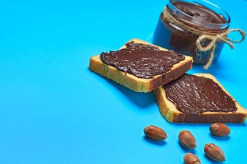 Glass jar full of melted chocolate or cream near pieces of vanilla bread and hazelnuts lies on blue desk on kitchen. Concept of nutritious breakfast or lunch