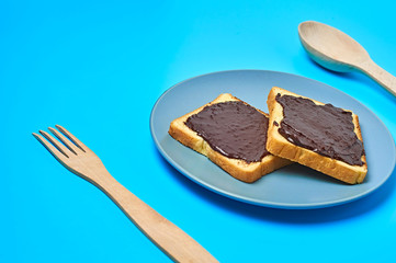 Two pieces of vanilla bread with melted chocolate on gray plate near wooden spoon and fork lies on blue desk on kitchen. Concept of nutritious breakfast or lunch