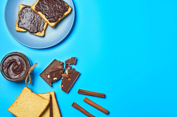 Glass jar full of melted chocolate near pieces of bread on plate and bamboo mat beside cinnamon sticks lies on blue desk on kitchen. Concept of nutritious breakfast or lunch