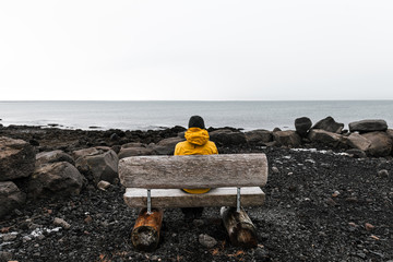 Lonely traveler man in yellow rain jacket  is relaxing at nice view of sea with wooden bench in Reykjavik, Iceland.