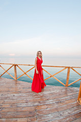 Charming girl in a long red dress is dancing and posing on the pier against the background of the...