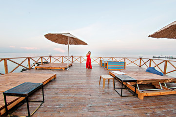 Charming girl in a long red dress is dancing and posing on the pier against the background of the sea.