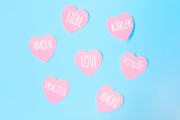 Some heart-shaped pink sticky notes with the word love written in different languages, such as English, Spanish, Italian, French, Dutch on a blue background. Valentines day, love concept.