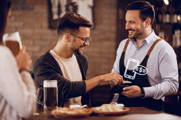 Young man making contactless payment to a waiter in a pub.