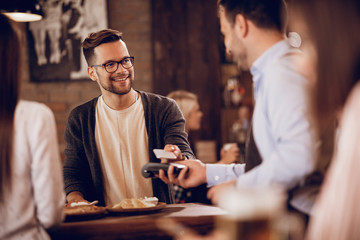 Happy man paying to waiter via contactless payment in a pub.