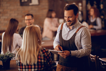 Happy waiter talking to a woman while writing her order on touchpad in a bar.