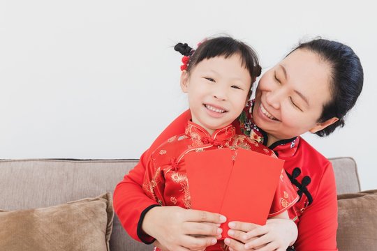 Smiling little Asian Chinese daughter receiving Chinese New Year red envelope packet from mother sitting on sofa at home with copyspace.Happy chinese new year family concept.