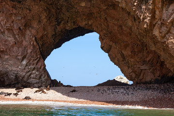 Large through grotto with a beach on which sea lions lie. Through the grotto, birds in the sky Ballestas Islands, Paracas reserv, Peru, Latin America.