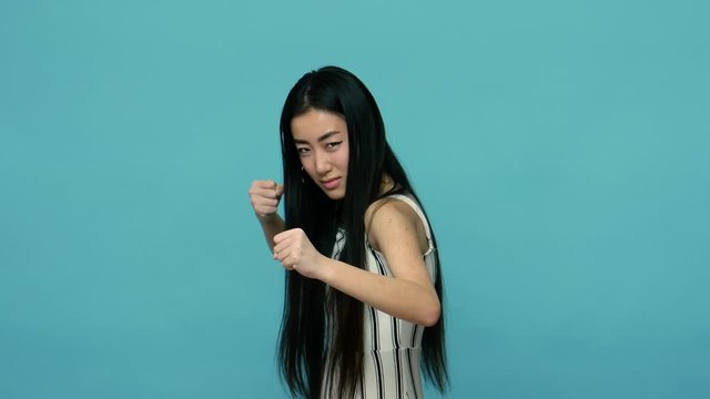 Let's fight! Aggressive asian woman with long straight black hair in dress ready to boxing with clenched fists, threatening to punch, fighting spirit. indoor studio shot isolated on blue background
