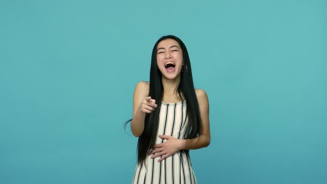 Extremely happy asian woman with long straight black hair in dress keeping hand on belly and laughing loudly and hysterically, pointing to camera, crazy laughter. indoor studio shot, blue background