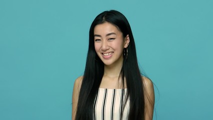 Beautiful and cunning asian woman with long straight black hair smiling slyly at camera and pondering tricky plan to cheat, do dirty tricks, prank someone. indoor studio shot, blue background