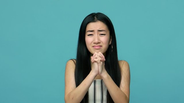 Please, forgive me! Unhappy asian woman with long straight black hair looking pleadingly at camera and holding cupped hands, praying, apologizing. indoor studio shot isolated on blue background