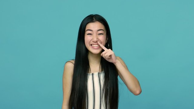 Funny Crazy Asian Woman With Long Black Hair Showing Tongue And Pulling Out Boogers, Picking Her Nose With Stupid Comical Expression, Making Faces. Indoor Studio Shot Isolated On Blue Background