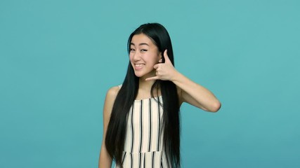 Hey you, call me! Attractive woman with long straight black hair showing talking on telephone gesture and pointing to camera, asking dial my number. indoor studio shot isolated on blue background