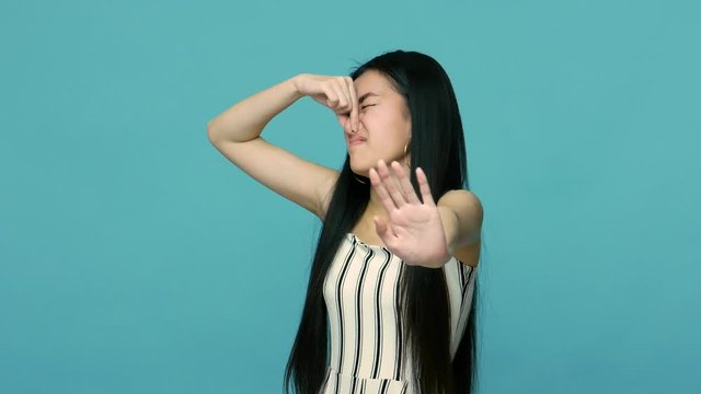 Stop bad smell! Displeased asian woman with long black hair pinching her nose and showing refusal gesture, grimacing in disgust, avoiding stinky breath. indoor studio shot isolated on blue background