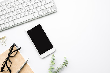 Smartphone with blank screen is on top of white office desk table with supplies. Top view with copy...
