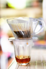 Drip brewing; clear glass of coffee mug and paper dripping bag with ground coffee in soft sunlight at home.