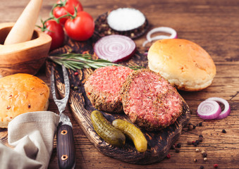 Fresh raw minced pepper beef burgers on vintage chopping board with buns onion and tomatoes on wooden background. Mortar with pestle.