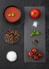 Black bowl plate of creamy tomato soup on black table background with stone chopping board and raw tomatoes, pepper and salt.