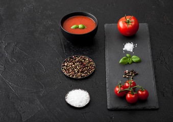 Black bowl plate of creamy tomato soup on black table background with stone chopping board and raw tomatoes, pepper and salt.