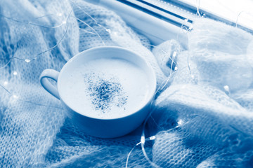 Cup with cappuccino on a warm scarf in the light of lights. Winter cozy life style, top view,toned classic blue color.
