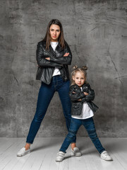 Positive young woman and little girl in similar jeans and black leather jackets standing in same pose over concrete background