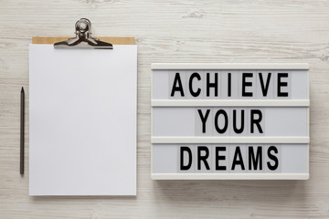 'Achieve your dreams' words on a lightbox, clipboard with blank sheet of paper on a white wooden surface, top view. Overhead, from above, flat lay.