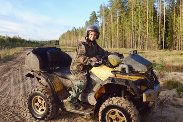 Fototapeta na wymiar Young beautiful tanned woman dressed in special clothes under disguise wears helmet with glasses rides powerful Quad bike with large studded wheels, girl smiles, looks happy. Against background forest