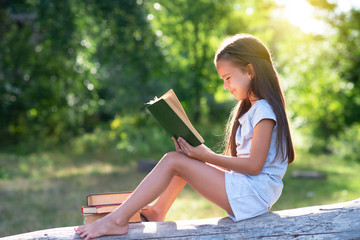 Charming little girl with long brown hair reads book outdoor sitting on tree in summer park or in a...