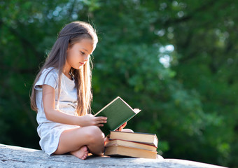 Obraz premium Charming little girl with long brown hair reads book outdoor sitting on tree in summer park or in a forest glade. Child booklover