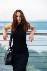 Fashion model wearing black elegant dress posing outdoor at the bank of Dniper river in Kyiv, Ukraine. Young beautiful brunette caucasian woman walking summer streets. Beautiful girl, urban portrait.