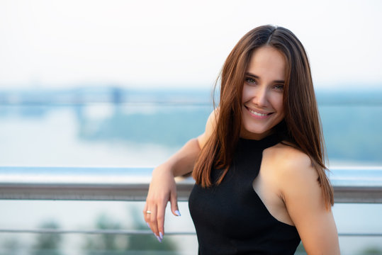 Fashion model wearing black elegant dress posing outdoor at the bank of Dniper river in Kyiv, Ukraine. Young beautiful brunette caucasian woman walking summer streets. Beautiful girl, urban portrait.