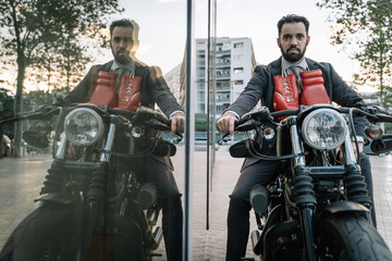 Businessman wearing a suit and touching boxing gloves sitting on a motorcycle next to a glass building where he is reflected