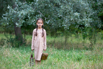 Pretty little girl with long pigtails and beautiful dirty face stands at summer meadow and holding vintage paraffin stove and flask. Orphan, child of war, poverty, hunger, famine, poor destitute kid.