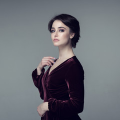 Fototapeta premium Beauty female portrait. Young attractive woman posing at studio over gray background. Beautiful model with perfect make-up dressed evening burgundy dress looking at camera.